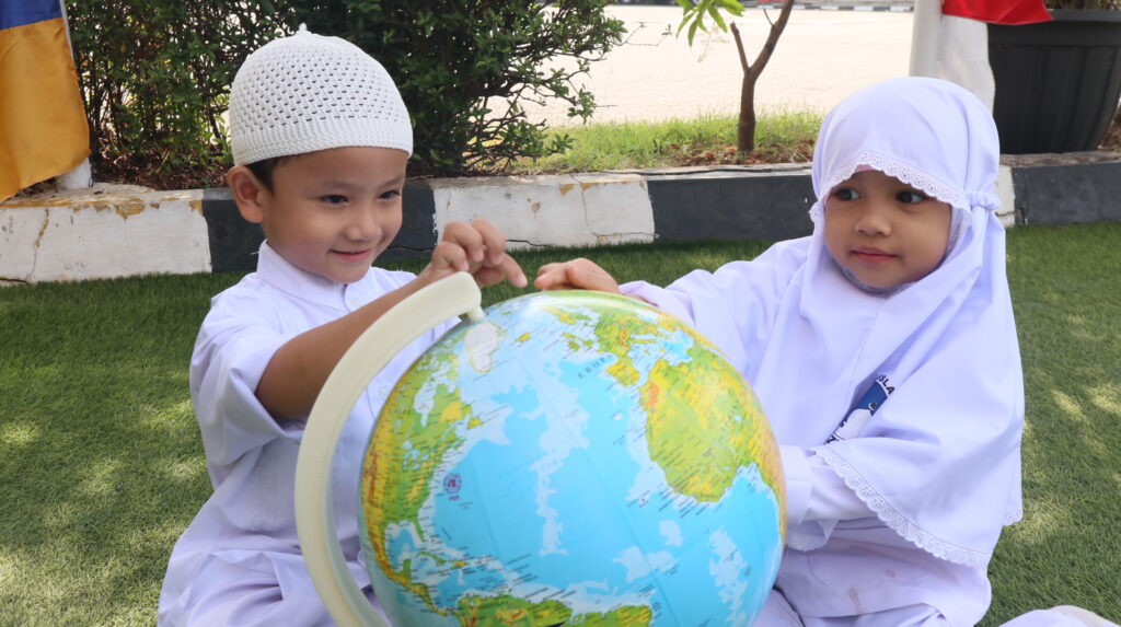 Dua murid TK Islam sedang belajar mengenal globe dan peta dunia di area outdoor sekolah.