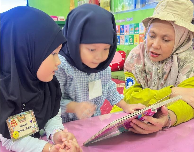 Seorang guru TK Islam Al Azhar 12 Cikarang membacakan buku cerita bergambar kepada dua orang murid perempuan.