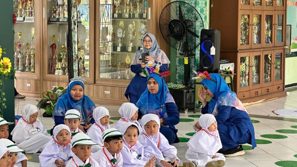 Momen keakraban guru yang mengenakan bando warna-warni bersama murid-murid baru yang duduk rapi di area sekolah.